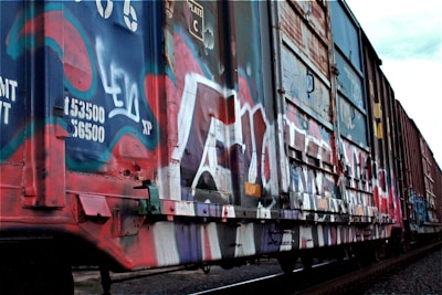 A freight train car covered in vibrant graffiti artwork is seen on a section of railway track. The graffiti features a mix of colors such as blue, red, and white, with various tags and designs painted over the entire side of the car. The surrounding environment appears industrial, and the train car shows signs of wear and tear.