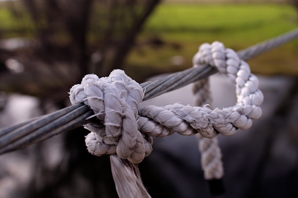 A close-up view of a thick, white rope tied in a knot around a metal cable against a blurred natural background with green and brown tones.