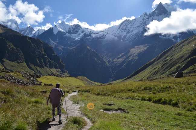 A hiker walks along a grassy trail surrounded by towering mountains, with snow-capped peaks and scattered fluffy clouds in the sky. The scene captures a stunning natural landscape bathed in sunlight, creating dramatic shadows on the slopes.