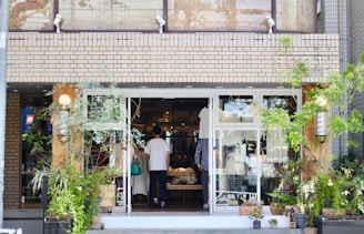 A small, charming shop with a brick exterior and large glass doors surrounded by lush green plants. Inside, a few people can be seen browsing items, creating a cozy and inviting atmosphere.
