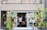 A small, charming shop with a brick exterior and large glass doors surrounded by lush green plants. Inside, a few people can be seen browsing items, creating a cozy and inviting atmosphere.