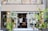 A small, charming shop with a brick exterior and large glass doors surrounded by lush green plants. Inside, a few people can be seen browsing items, creating a cozy and inviting atmosphere.