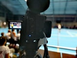 A camera on a tripod is prominently displayed with its viewfinder screen showing footage of an indoor sports event. The background is an indoor sports hall with a blue floor, and numerous spectators are faintly visible.