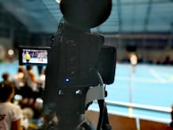 A camera on a tripod is prominently displayed with its viewfinder screen showing footage of an indoor sports event. The background is an indoor sports hall with a blue floor, and numerous spectators are faintly visible.