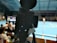 A camera on a tripod is prominently displayed with its viewfinder screen showing footage of an indoor sports event. The background is an indoor sports hall with a blue floor, and numerous spectators are faintly visible.