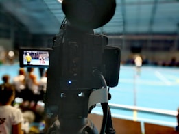 A camera on a tripod is prominently displayed with its viewfinder screen showing footage of an indoor sports event. The background is an indoor sports hall with a blue floor, and numerous spectators are faintly visible.