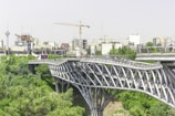 A modern, multi-level pedestrian bridge with a distinctive lattice structure spans over a green park area. Below and surrounding the bridge, lush trees and foliage create a natural environment. In the background, a cityscape with numerous buildings is visible, along with construction cranes and a tall tower.