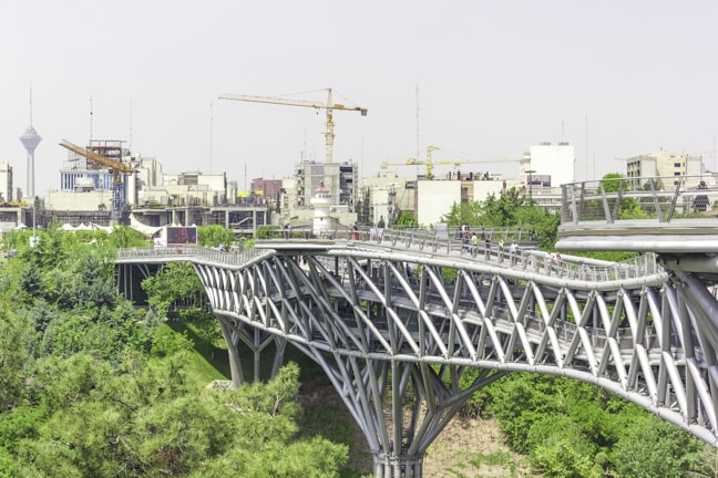 A modern, multi-level pedestrian bridge with a distinctive lattice structure spans over a green park area. Below and surrounding the bridge, lush trees and foliage create a natural environment. In the background, a cityscape with numerous buildings is visible, along with construction cranes and a tall tower.