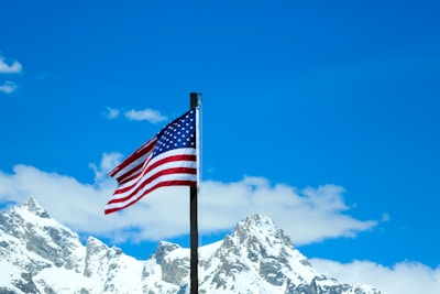 An American flag waves prominently in the foreground, set against a background of snow-covered mountain peaks under a clear blue sky. The contrast between the vibrant colors of the flag and the serene natural landscape creates a striking visual.