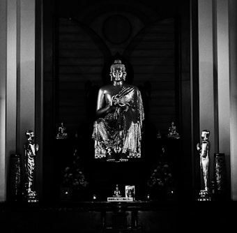 A prominent statue of the Buddha is centrally placed in a dark shrine, flanked by two smaller metallic statues in a reverent posture. The setting conveys a sense of reverence and peace, emphasized by the subdued lighting that highlights the intricate details of the main statue's garment.