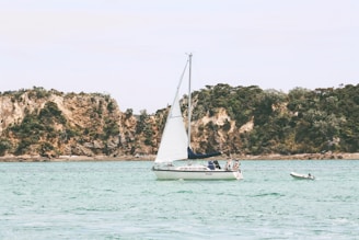 A sailboat with white sails is calmly floating on clear, turquoise waters. Two people are visible on the boat, appearing relaxed. In the background, rugged, tree-covered cliffs rise, providing a serene and natural backdrop. A smaller dinghy is tethered nearby.