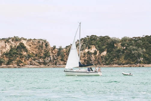 A sailboat with white sails is calmly floating on clear, turquoise waters. Two people are visible on the boat, appearing relaxed. In the background, rugged, tree-covered cliffs rise, providing a serene and natural backdrop. A smaller dinghy is tethered nearby.