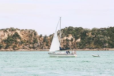 A sailboat with white sails is calmly floating on clear, turquoise waters. Two people are visible on the boat, appearing relaxed. In the background, rugged, tree-covered cliffs rise, providing a serene and natural backdrop. A smaller dinghy is tethered nearby.