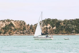 A sailboat with white sails is calmly floating on clear, turquoise waters. Two people are visible on the boat, appearing relaxed. In the background, rugged, tree-covered cliffs rise, providing a serene and natural backdrop. A smaller dinghy is tethered nearby.