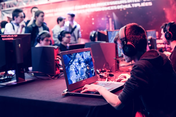 Several people are seated and focused on computer screens, wearing headsets, in an environment that appears to be a gaming tournament or esports event. The room is dimly lit with neon lighting, highlighting screens and creating an intense atmosphere.