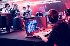 Several people are seated and focused on computer screens, wearing headsets, in an environment that appears to be a gaming tournament or esports event. The room is dimly lit with neon lighting, highlighting screens and creating an intense atmosphere.