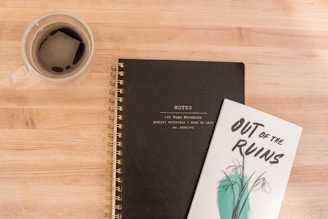 A wooden surface hosts a spiral-bound notebook with the word 'Notes' embossed on its black cover. Adjacent to it lies a book titled 'Out of the Ruins' featuring an abstract plant design. A transparent glass mug, filled with coffee, stands to the left of the notebooks.