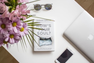 A neatly arranged white desk surface displays a book titled 'How's Your Soul?' with a pair of sunglasses resting above it, an open silver laptop on the right, and a smartphone placed at the bottom. Pink flowers with green stems are seen in the upper left corner, adding a vibrant touch to the minimalist setting.