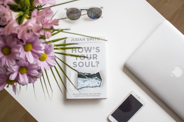A neatly arranged white desk surface displays a book titled 'How's Your Soul?' with a pair of sunglasses resting above it, an open silver laptop on the right, and a smartphone placed at the bottom. Pink flowers with green stems are seen in the upper left corner, adding a vibrant touch to the minimalist setting.