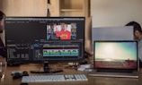 Two computer screens on a desk, one displaying video editing software with a timeline and footage of a person in a gym wearing a red hoodie. The other screen shows a serene beach scene with a surfer walking towards the ocean. A keyboard, a computer mouse, sunglasses, and a glass of water are visible on the desk. In the background, parts of two people are visible, working on computers.