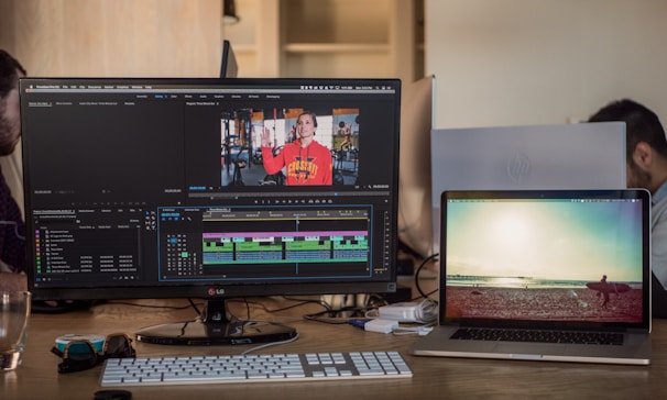 Two computer screens on a desk, one displaying video editing software with a timeline and footage of a person in a gym wearing a red hoodie. The other screen shows a serene beach scene with a surfer walking towards the ocean. A keyboard, a computer mouse, sunglasses, and a glass of water are visible on the desk. In the background, parts of two people are visible, working on computers.