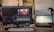 Two computer screens on a desk, one displaying video editing software with a timeline and footage of a person in a gym wearing a red hoodie. The other screen shows a serene beach scene with a surfer walking towards the ocean. A keyboard, a computer mouse, sunglasses, and a glass of water are visible on the desk. In the background, parts of two people are visible, working on computers.