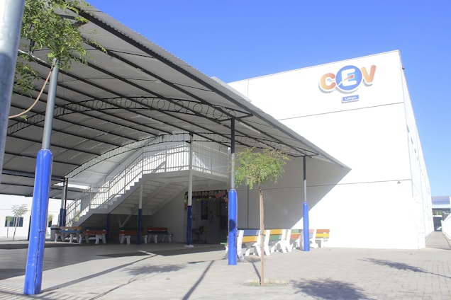 A modern, covered outdoor area with a white metal staircase leading to an elevated walkway. The structure is supported by blue pillars, and there are picnic-style benches with colorful backs underneath. A young tree grows near the center, casting shadows on the paved ground. The building has a logo with large, colorful letters on its facade.