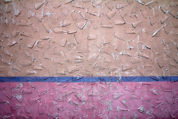 The image features a textured wall with abstract patterns. The upper portion of the wall is painted in a beige color, while the lower portion is pink, separated by a thin blue line. The surface has rough, uneven protrusions giving it a rustic, unfinished look.
