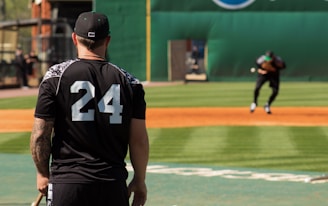 A person wearing a black jersey with the number 24 is standing on a baseball field, observing a player in the distance who is practicing or playing. The setting is a sunny day with a well-maintained field and a green backdrop.