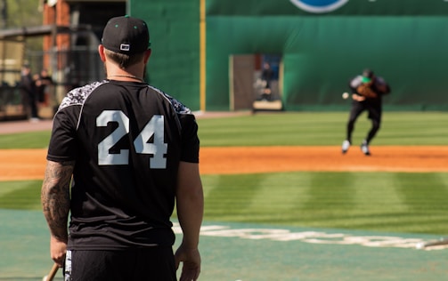 A person wearing a black jersey with the number 24 is standing on a baseball field, observing a player in the distance who is practicing or playing. The setting is a sunny day with a well-maintained field and a green backdrop.