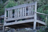 A weathered wooden bench with a simple design, situated outdoors against a backdrop of lush greenery and rocks. The bench is elevated on a slight incline, giving it an aged and rustic appearance surrounded by natural elements.