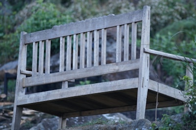 A weathered wooden bench with a simple design, situated outdoors against a backdrop of lush greenery and rocks. The bench is elevated on a slight incline, giving it an aged and rustic appearance surrounded by natural elements.