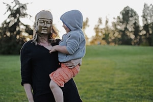 woman with mask carrying toddler with hoodie on grass field