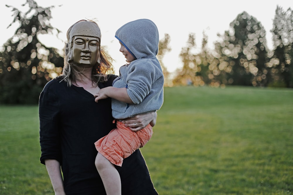 Femme avec un masque portant un enfant en bas âge avec un sweat à capuche sur un terrain en herbe