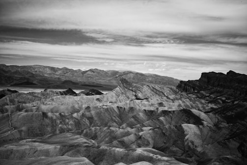 A black and white photograph of a rugged, mountainous landscape with barren, layered rock formations. The sky is cloudy with a mix of light and dark shades, adding depth and contrast to the scene. The distant mountains appear hazy, creating a sense of vastness.
