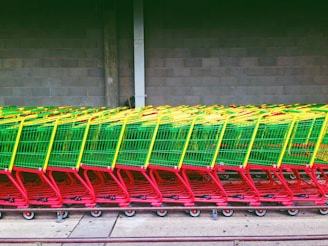 A row of colorful shopping carts is neatly lined up against a concrete wall, featuring a vibrant combination of green baskets with yellow handles and red frames. The carts are on a sidewalk, and the setting seems to be an outdoor area near a store or warehouse.