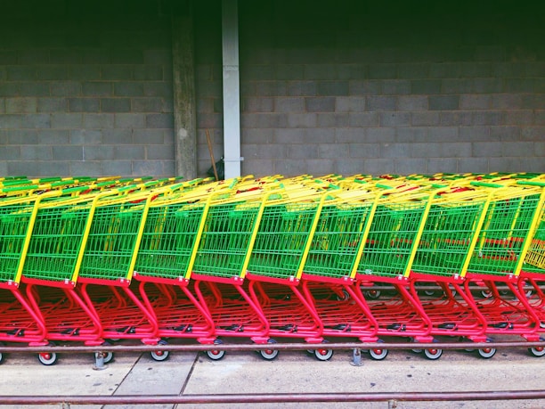 A row of colorful shopping carts is neatly lined up against a concrete wall, featuring a vibrant combination of green baskets with yellow handles and red frames. The carts are on a sidewalk, and the setting seems to be an outdoor area near a store or warehouse.