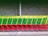 A row of colorful shopping carts is neatly lined up against a concrete wall, featuring a vibrant combination of green baskets with yellow handles and red frames. The carts are on a sidewalk, and the setting seems to be an outdoor area near a store or warehouse.