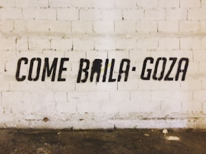 Black stencil graffiti on a textured white brick wall spells out the phrase 'COME BAILA GOZA'. The floor is worn and has various stains.