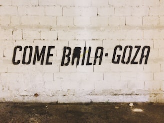 Black stencil graffiti on a textured white brick wall spells out the phrase 'COME BAILA GOZA'. The floor is worn and has various stains.