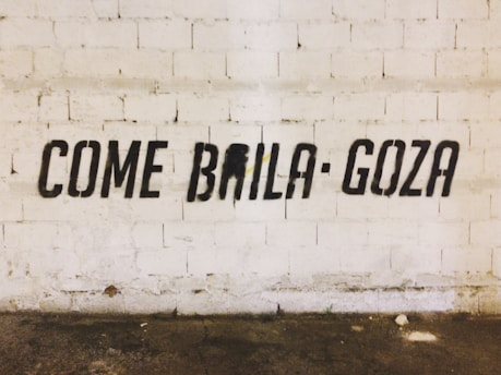 Black stencil graffiti on a textured white brick wall spells out the phrase 'COME BAILA GOZA'. The floor is worn and has various stains.
