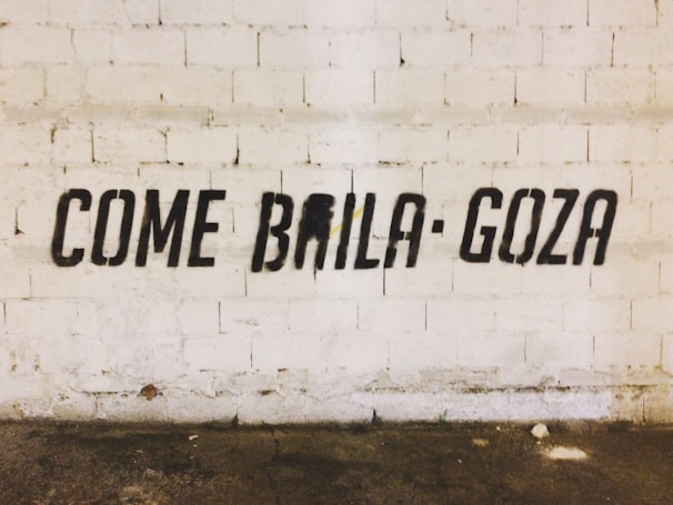 Black stencil graffiti on a textured white brick wall spells out the phrase 'COME BAILA GOZA'. The floor is worn and has various stains.