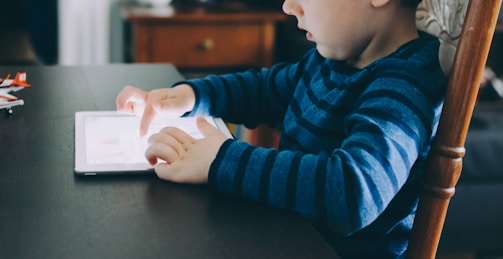 A young child is sitting at a table, engaging with a tablet device using their hands. The child is wearing a long-sleeved blue striped shirt and is seated in a wooden chair. On the table, some small toy airplanes are visible in the background.