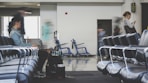 A spacious airport lounge with rows of empty blue chairs. A few people are present, with one person sitting and another standing while looking at a phone. There is movement indicating some are in transit, with blurred figures suggesting motion. Luggage and a wheelchair are visible in the background, hinting at a travel setting.