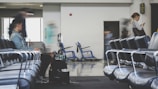 A spacious airport lounge with rows of empty blue chairs. A few people are present, with one person sitting and another standing while looking at a phone. There is movement indicating some are in transit, with blurred figures suggesting motion. Luggage and a wheelchair are visible in the background, hinting at a travel setting.