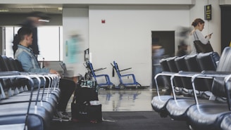 A spacious airport lounge with rows of empty blue chairs. A few people are present, with one person sitting and another standing while looking at a phone. There is movement indicating some are in transit, with blurred figures suggesting motion. Luggage and a wheelchair are visible in the background, hinting at a travel setting.