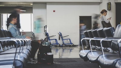 A spacious airport lounge with rows of empty blue chairs. A few people are present, with one person sitting and another standing while looking at a phone. There is movement indicating some are in transit, with blurred figures suggesting motion. Luggage and a wheelchair are visible in the background, hinting at a travel setting.