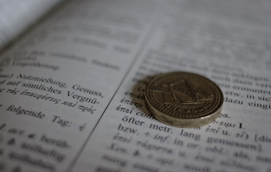 A coin is resting on an open book, with text in both German and Greek visible on the pages. The focus on the coin creates a blurred background effect on the text.
