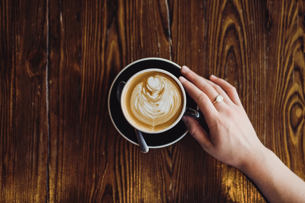 person holding cappuccino in teapot with saucer on table