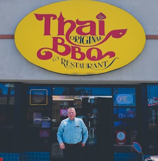 A man stands in front of a Thai BBQ restaurant. The entrance features a bright yellow sign with red lettering above the door. The man is wearing a light blue shirt and dark pants, posed directly under the sign, with one hand in his pocket. Neon signs and posters are visible on the glass windows and door.